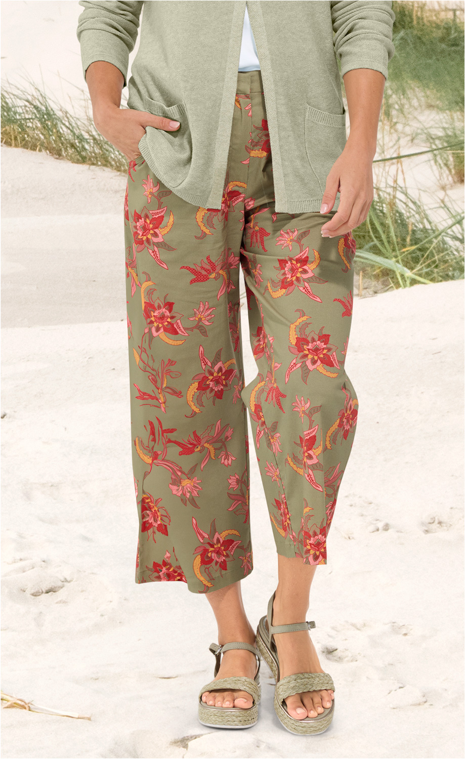 Baumwoll-Mix-Hose in khaki mit weitem Bein und floralem Muster in rot, pink und gelb, kombiniert mit grünem Strickcardigan, Pop-up-Sandalen mit geflochtenem Riemen, Strand und Dünen im Hintergrund