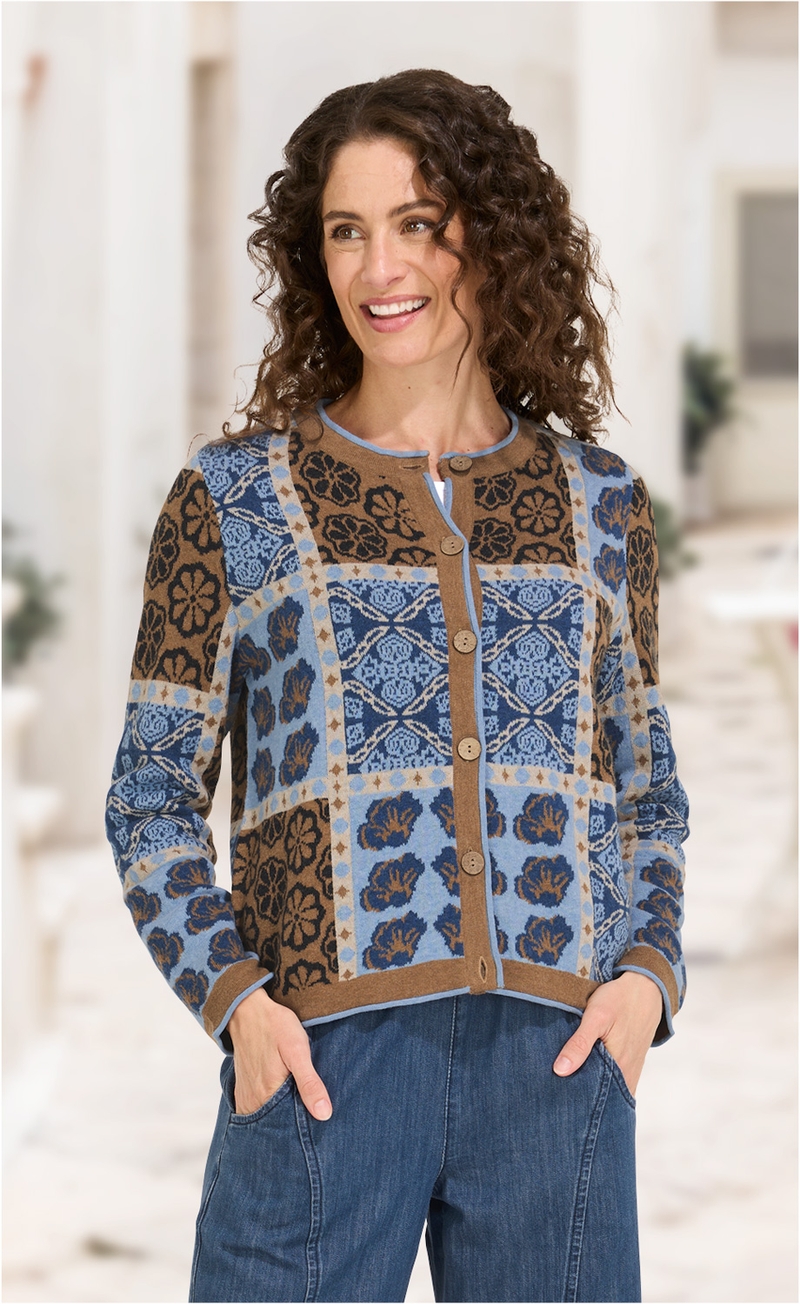 Frau trägt Baumwoll-Mix-Jacke mit blau-braunem floralen Karomuster, Knopfleiste und Rundhalsausschnitt, kombiniert mit blauen Jeans, Hände in den Taschen, lockige braune Haare, lächelnd, unscharfer heller Hintergrund.