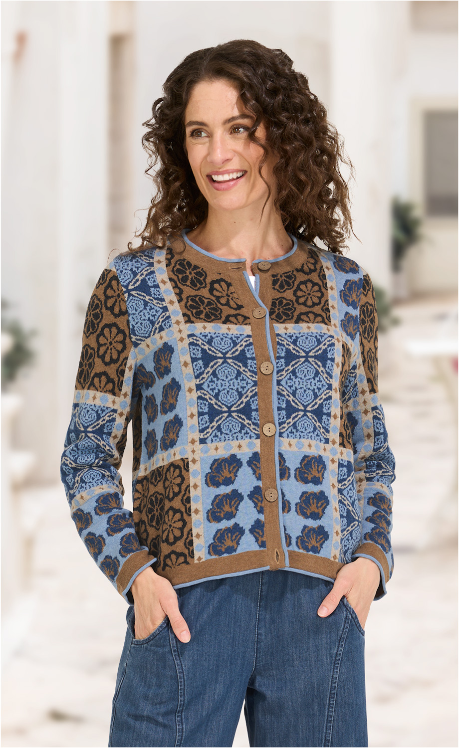Frau trägt Baumwoll-Mix-Jacke mit blau-braunem floralen Karomuster, Knopfleiste und Rundhalsausschnitt, kombiniert mit blauen Jeans, Hände in den Taschen, lockige braune Haare, lächelnd, unscharfer heller Hintergrund.