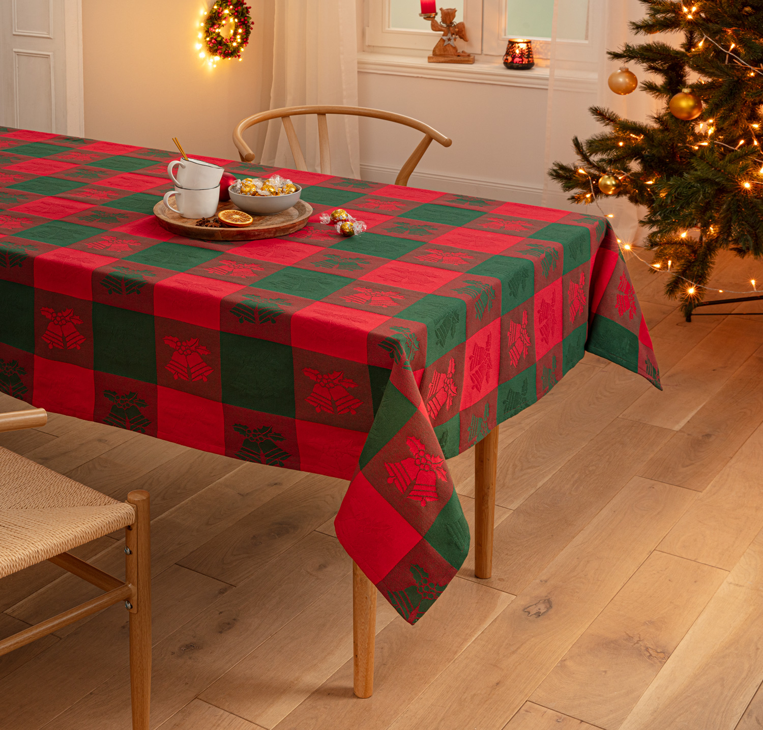 Baumwoll-Tischdecke 90x90 cm mit kariertem Weihnachtsdesign in Rot und Grün, Glockenmotiv, auf Holztisch, dekoriert mit zwei Tassen, Schale mit Pralinen, Weihnachtsbaum und festlichen Lichtern im Hintergrund