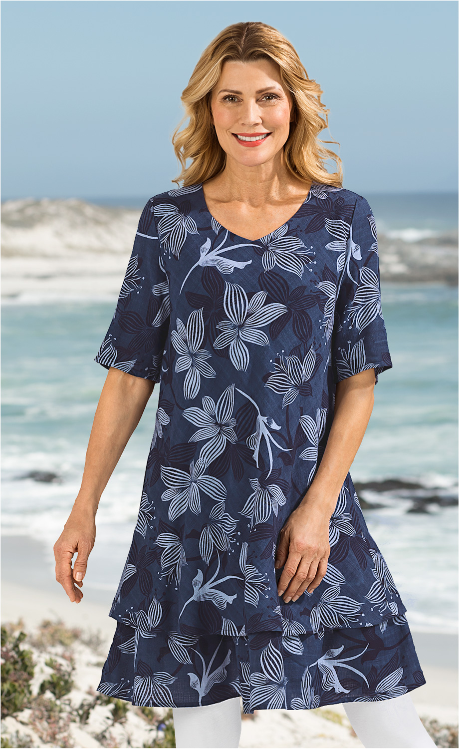 Frau am Strand trägt blaues Baumwoll-Mix-Kleid mit V-Ausschnitt und hellblauem Blumenmuster, kurze Ärmel, leichte Doppel-Lagen-Optik, kombiniert mit weißen Leggings, blonder welliger mittellanger Haarstil, lächelnd