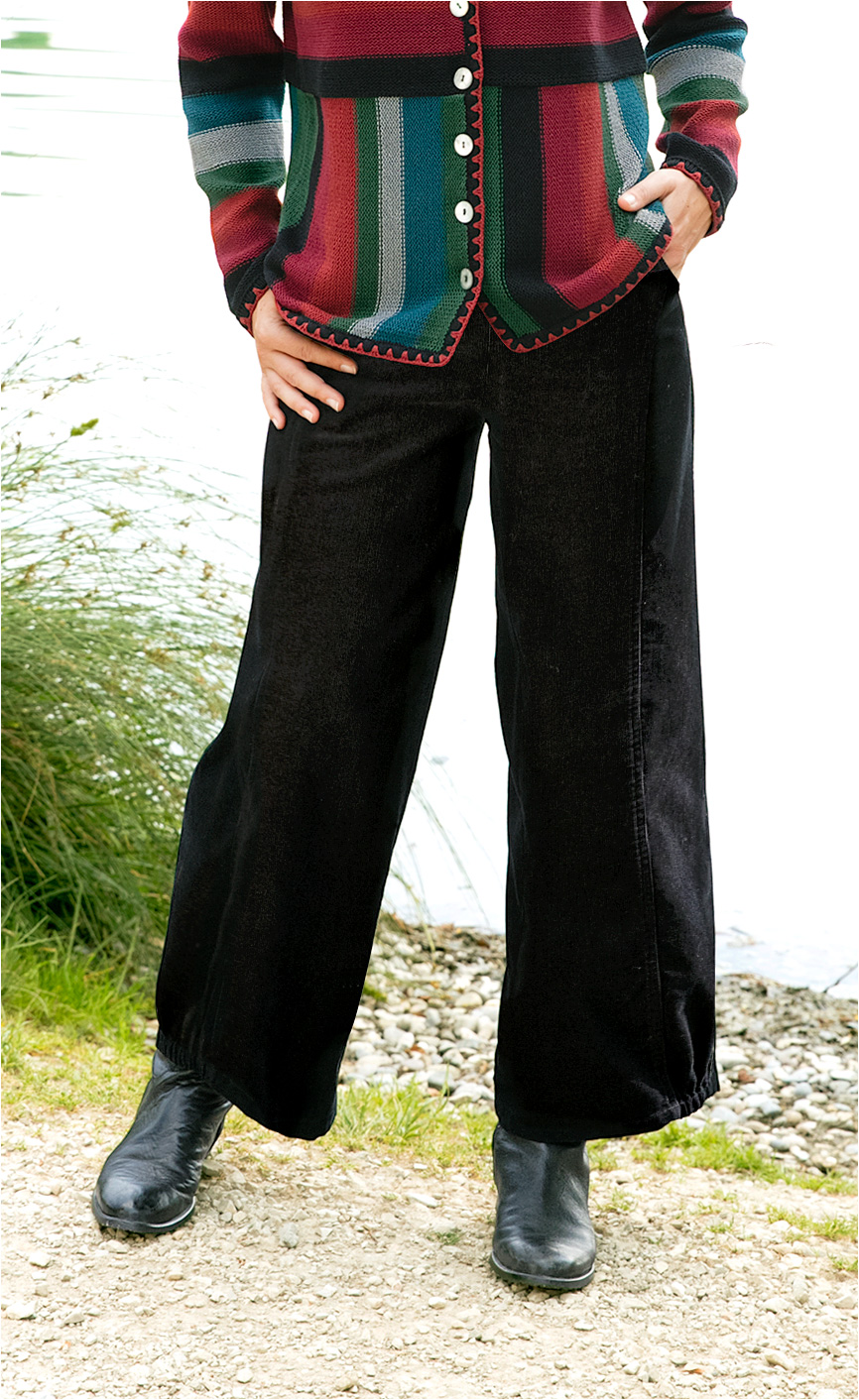 Baumwoll-Hose in Schwarz, weit geschnitten, mit elastischem Saum, kombiniert mit buntem, gestreiftem Wolljackett und schwarzen Lederstiefeln, stehend auf einem Kiesweg mit Gras und Wasser im Hintergrund.