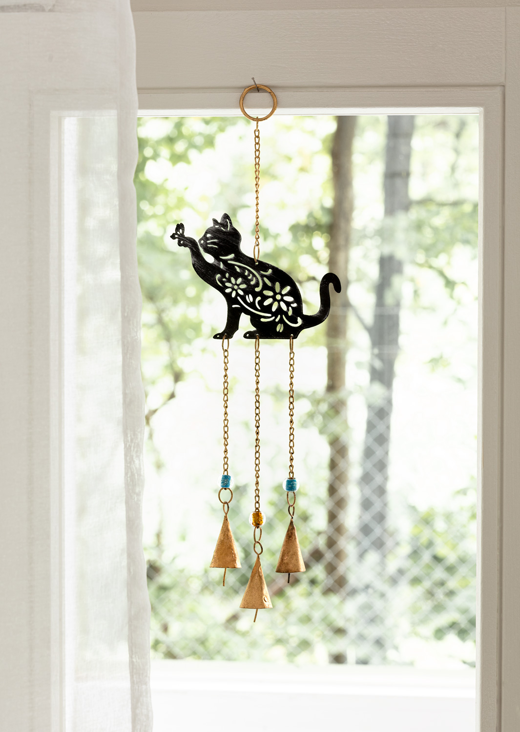 Metall Mobile mit schwarzer Katzenform, floral verziert, hängend an einer goldfarbenen Kette, darunter drei goldfarbene Glocken, zwei mit blauen und eine mit gelber Perle, im Hintergrund Fenster mit Sicht auf grüne Bäume und weißem Vorhang