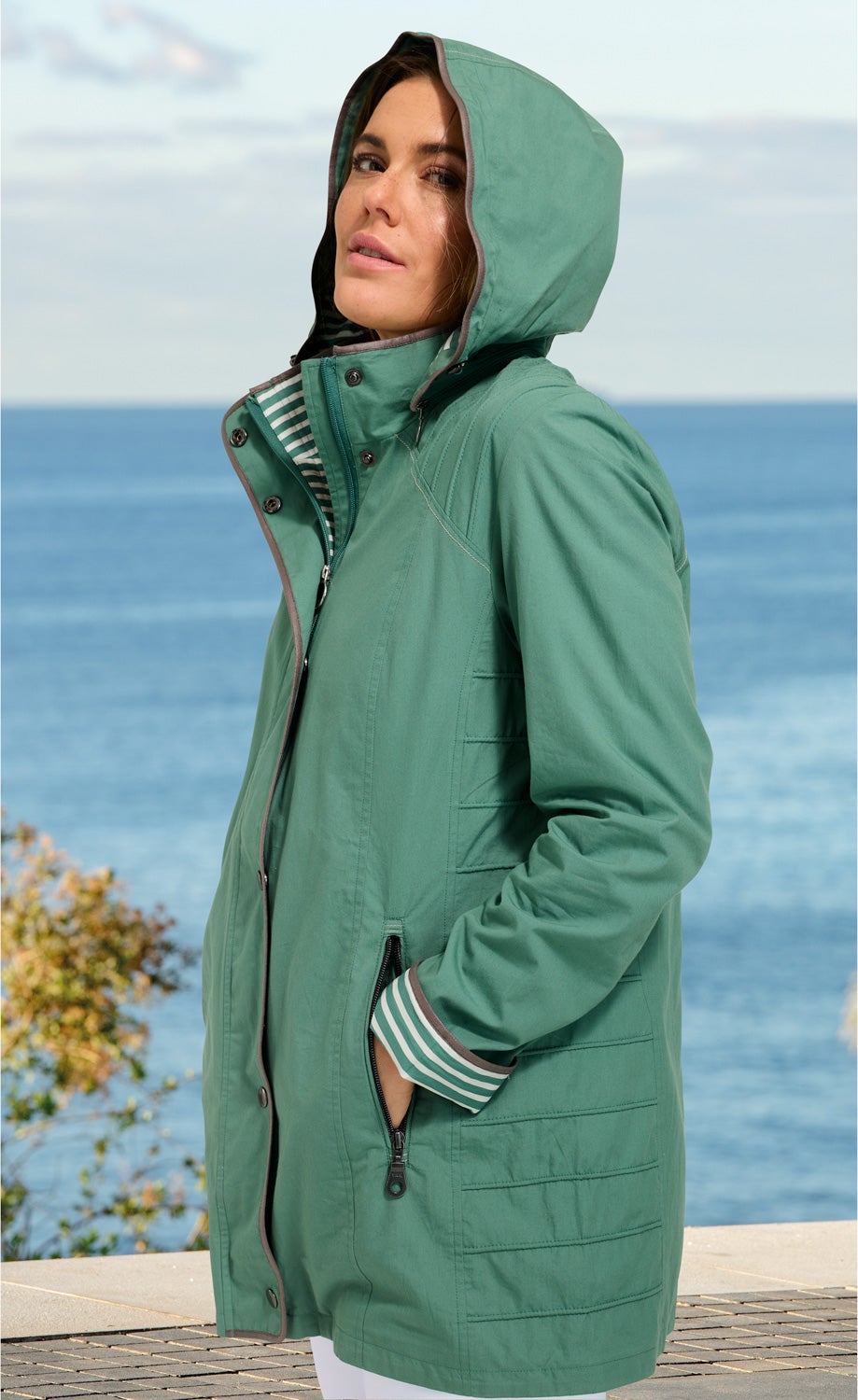 Frau trägt grüne Baumwoll-Jacke mit Kapuze und Reißverschlusstaschen, gestreifte Armbündchen, steht seitlich vor Meer und Himmel im Hintergrund