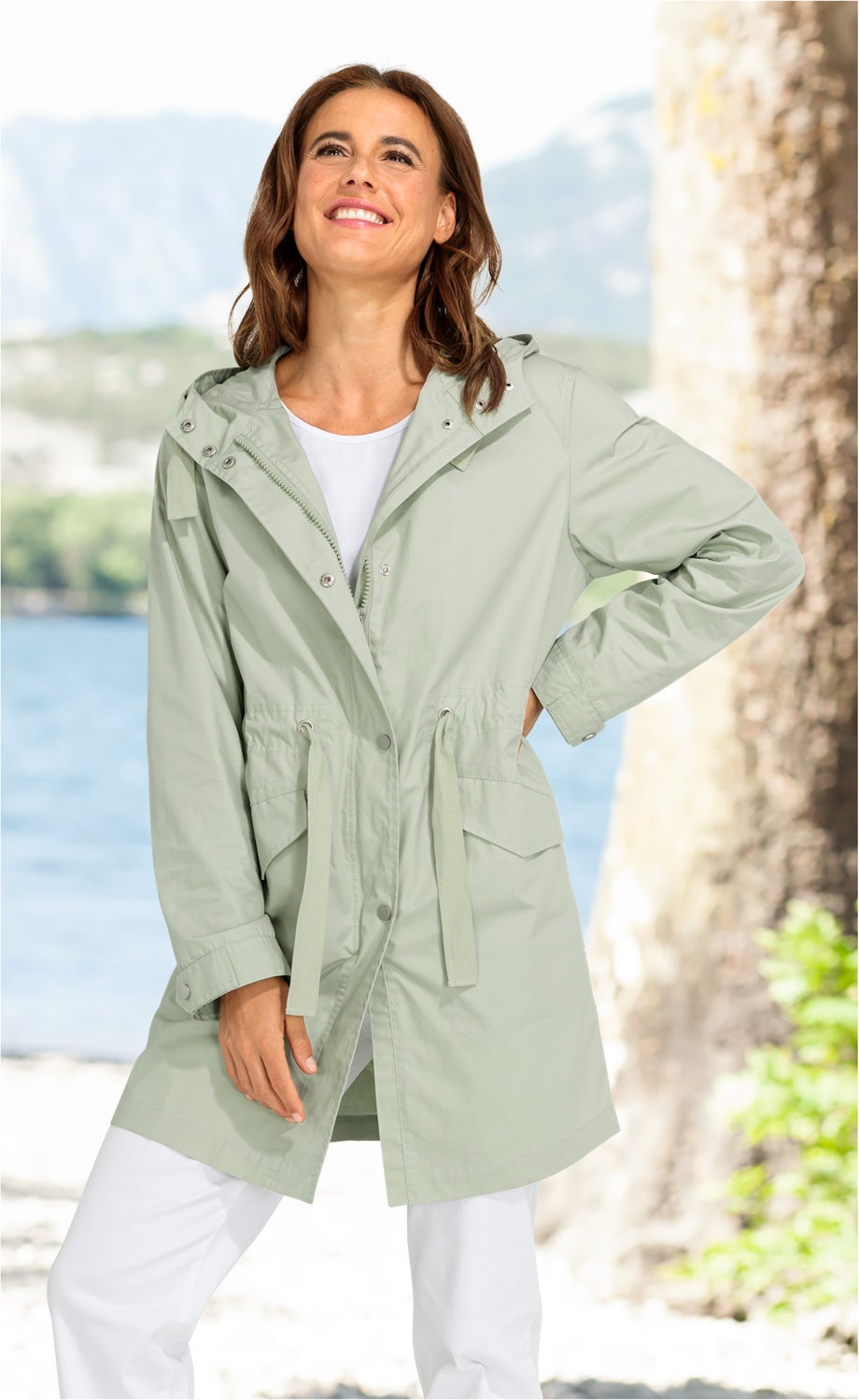 Frau trägt helle Baumwoll-Jacke mit Kapuze und Tunnelzug in der Taille, weißes Shirt und weiße Hose, steht lächelnd im Freien bei sonnigem Wetter vor verschwommenem See und Bäumen im Hintergrund