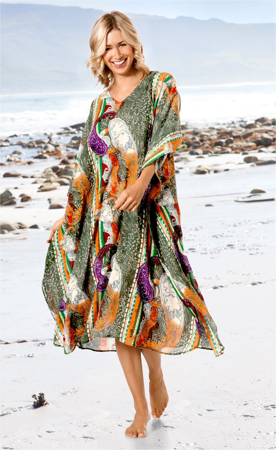 Frau am Strand trägt farbenfrohen Viskose-Kaftan mit Paisley- und Streifenmuster in Grün, Orange, Lila und Weiß, barfuß, lächelnd, mit blonden Haaren und lockeren Wellen, weißer Sand und Meer im Hintergrund