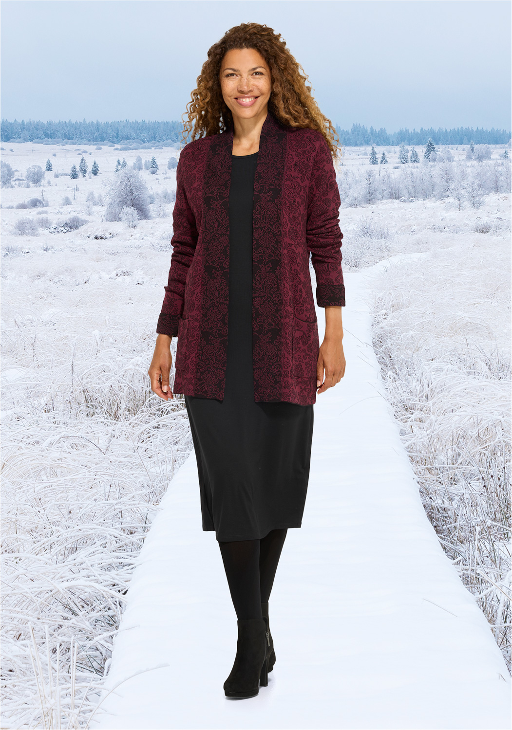 Frau mit langen lockigen Haaren trägt eine bordeauxrote Baumwoll-Strickjacke mit floralem Muster über einem schwarzen Kleid, schwarze Strumpfhose und schwarze Stiefeletten, stehend auf einem schneebedeckten Weg im Winterlandschaftshintergrund