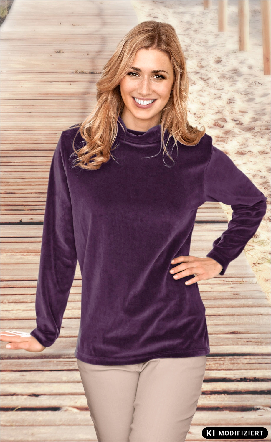 Frau mit langen blonden Haaren trägt ein violettes Baumwoll-Shirt mit hohem Kragen und langen Ärmeln, steht vor einem Holzsteg mit Sand im Hintergrund, lächelt, rechte Hand in Hüfte, linke Hand gestützt auf Holzgeländer