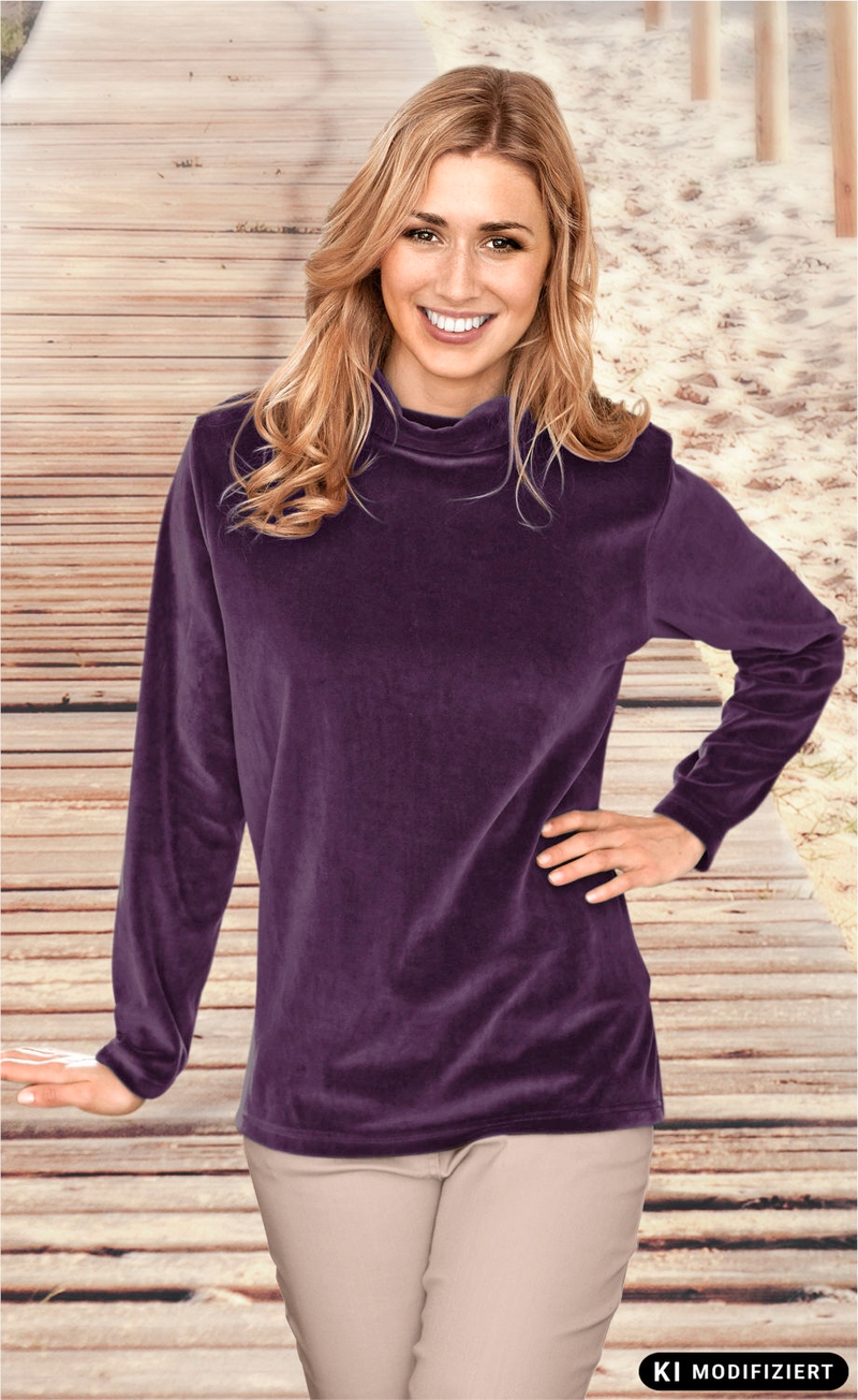 Frau mit langen blonden Haaren trägt ein violettes Baumwoll-Shirt mit hohem Kragen und langen Ärmeln, steht vor einem Holzsteg mit Sand im Hintergrund, lächelt, rechte Hand in Hüfte, linke Hand gestützt auf Holzgeländer