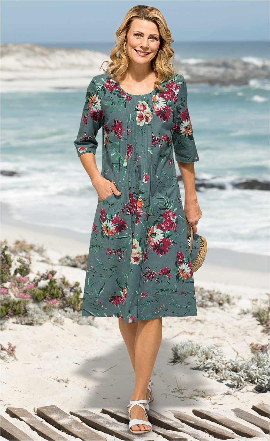Frau am Strand trägt grünes Baumwoll-Kleid mit buntem Blumenmuster, 3/4-Ärmel, Taschen, weiße Sandalen, hält Strohhandtasche, steht auf Holzsteg mit Sand, Meer und Felsen im Hintergrund, sonniger Tag