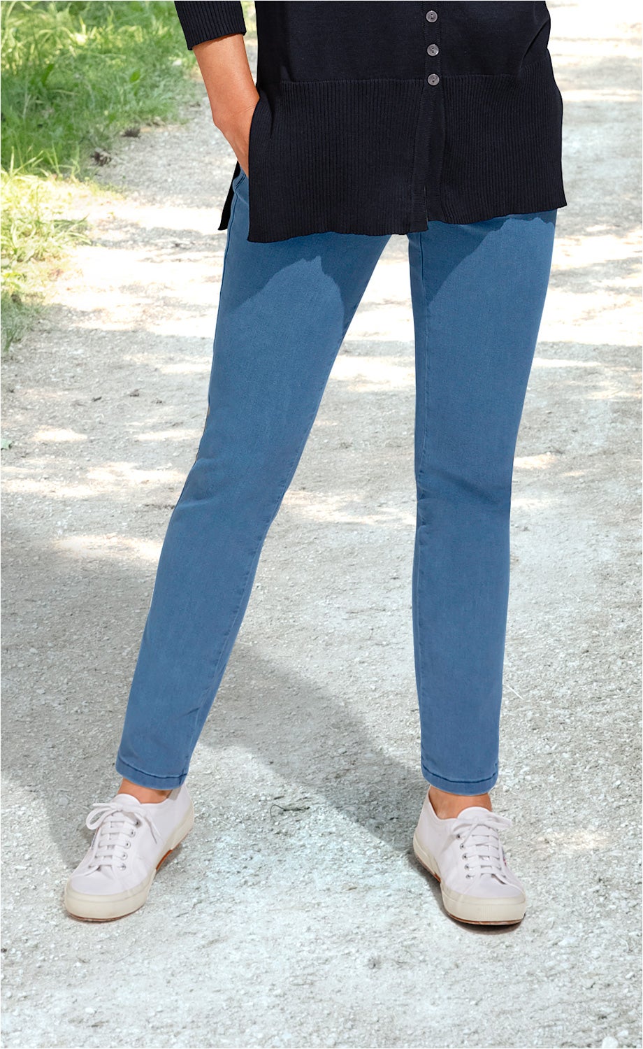 Frau trägt hellblaue Baumwoll-Mix-Hose mit schmalem Schnitt, dazu weiße Sneaker und schwarzen langen Pullover, im Freien auf einem Kiesweg stehend
