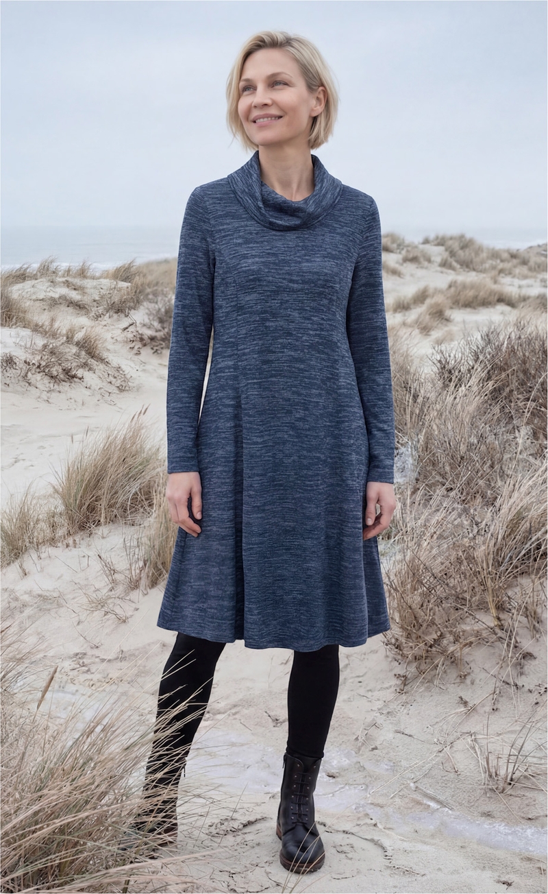 Frau mit kurzem blondem Haar trägt blaues, langärmeliges Kleid mit hohem Kragen, schwarze Leggings und schwarze Schnürstiefel, steht lächelnd am sandigen Dünenstrand mit trockenem Gras und bewölktem Himmel im Hintergrund