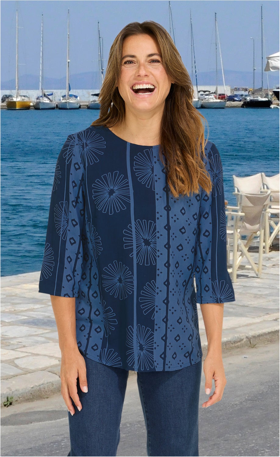 Frau mit langen braunen Haaren trägt blaue Viskose-Bluse mit floralem und geometrischem Muster, 3/4-Ärmeln, lächelt, steht vor Meer mit Segelbooten und Steg im Hintergrund