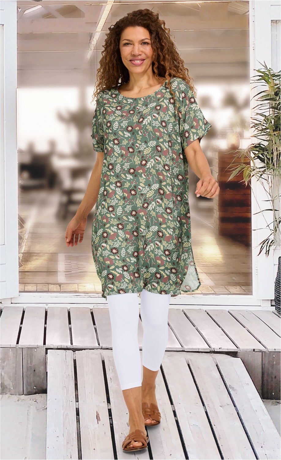 Frau trägt grün gemusterten Viskose-Kaftan mit floralem Design, kurze Ärmel, seitliche Schlitze, weiße Leggings und braune Sandalen, stehend vor hellem, modernem Innenraum mit Holzboden