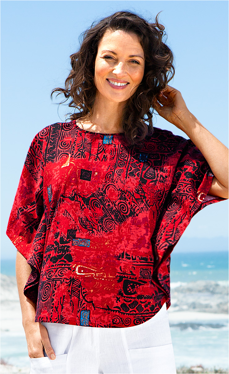 Lächende Frau am Meer trägt rotes, kurzärmeliges Baumwoll-Shirt mit abstraktem schwarzen und blauen Muster, kombiniert mit weißen Hosen, hellblauer Himmel im Hintergrund