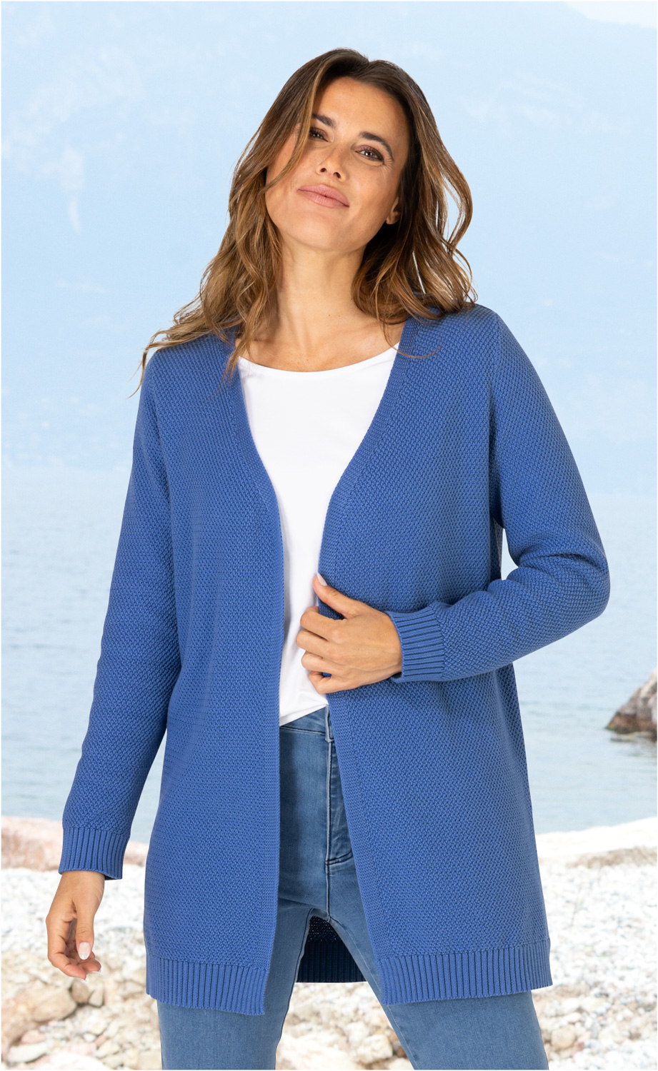 Frau trägt offene blaue Baumwoll-Strickjacke mit gerippten Bündchen, weiße Bluse und blaue Jeans, steht am Kiesstrand vor See und bewölktem Himmel