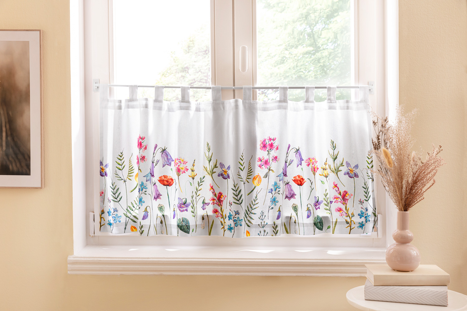 Weiße Küchengardine mit buntem Blumenmuster, hängend an einer Stange vor einem hellen Fenster, umgeben von beiger Wand, links ein gerahmtes Bild, rechts eine Vase mit Trockenblumen auf zwei Büchern aufgeräumt auf einem kleinen runden Tisch
