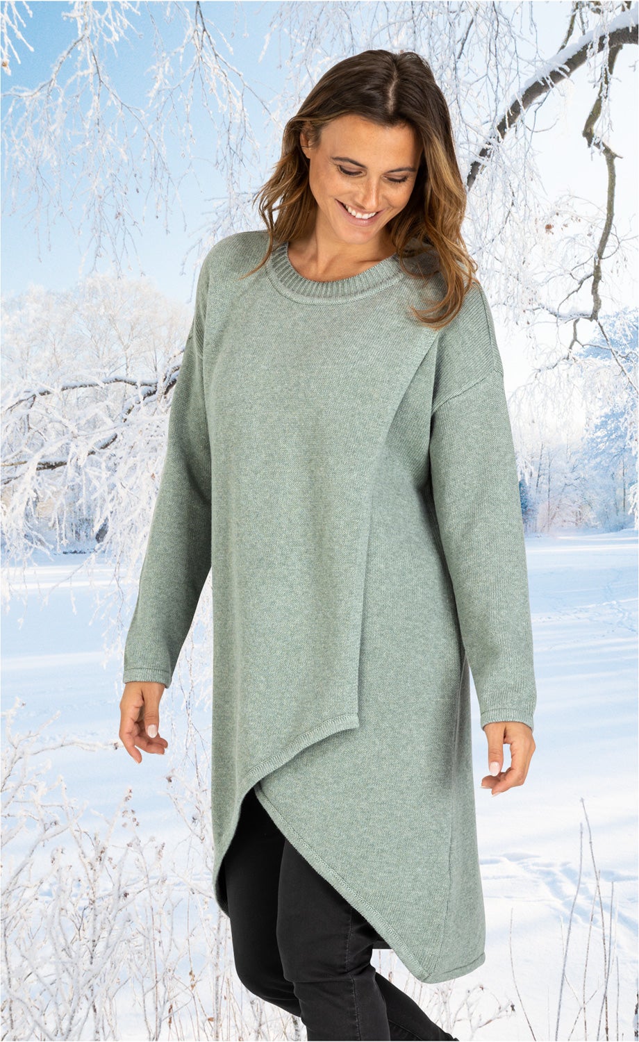 Frau trägt mintgrünes, asymmetrisches Baumwoll-Mix-Kleid mit langem Arm, stehend im verschneiten Winterwald, lächelnd, lockiges schulterlanges Haar, schwarze Hose sichtbar unter dem Kleid, frostiger Himmel und vereiste Bäume im Hintergrund.