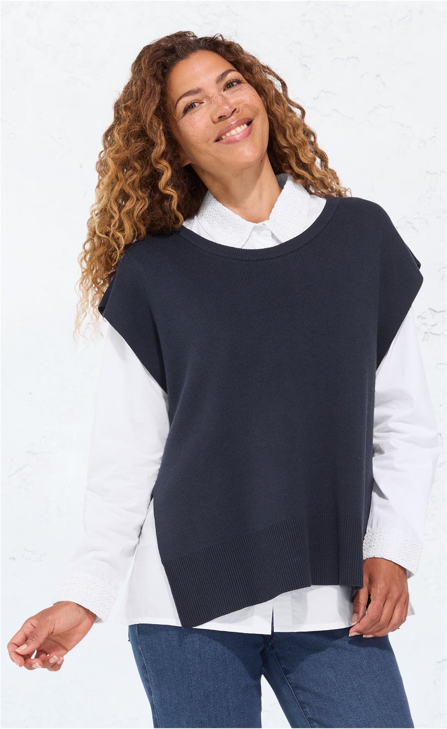 Frau mit lockigem Haar trägt weißen Langarmbluse und dunkelblauen Baumwoll-Mix-Pullunder, stehend vor hellem Hintergrund, lächelnd, casual Outfit mit Jeans