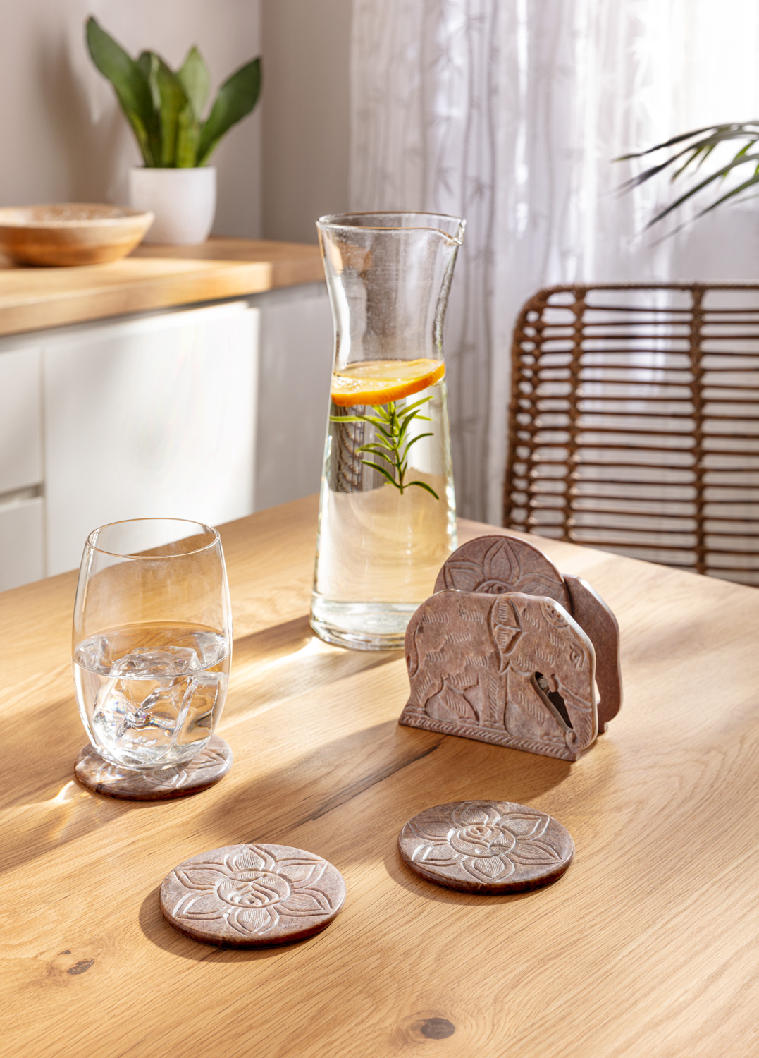 Set aus fünf braunen Untersetzern mit floralen und Elefanten-Motiven auf hellem Holztisch, Glas mit Eiswürfeln und Karaffe mit Wasser, Orangenscheibe und Rosmarinzweig, heller Raum mit Pflanzen und natürlichem Licht im Hintergrund
