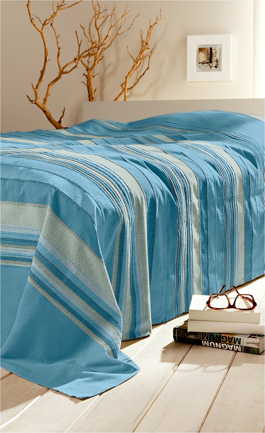 Baumwoll-Überwurf 250x180 in Blau mit gestreiftem Muster auf hellem Holzboden, dekorativ drapiert, im Hintergrund weiße Wand mit abstraktem Bild und Zweigen, neben dem Überwurf Bücherstapel mit brauner Brille darauf