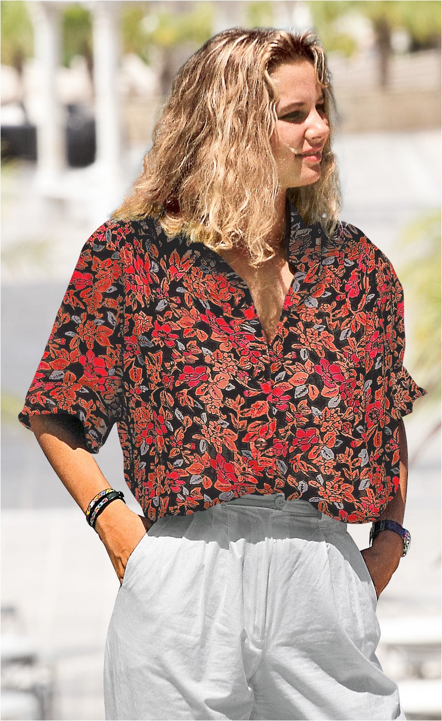 Frau trägt kurze Viskose-Mix-Bluse mit rotem und orangem Blumenmuster auf schwarzem Grund, halb in weiße Hose gesteckt, Arme in Taschen, blonder gewellter kinnlanger Haaransatz, Armbänder und Armbanduhr sichtbar, unscharfer Außenbereich im Hintergrund