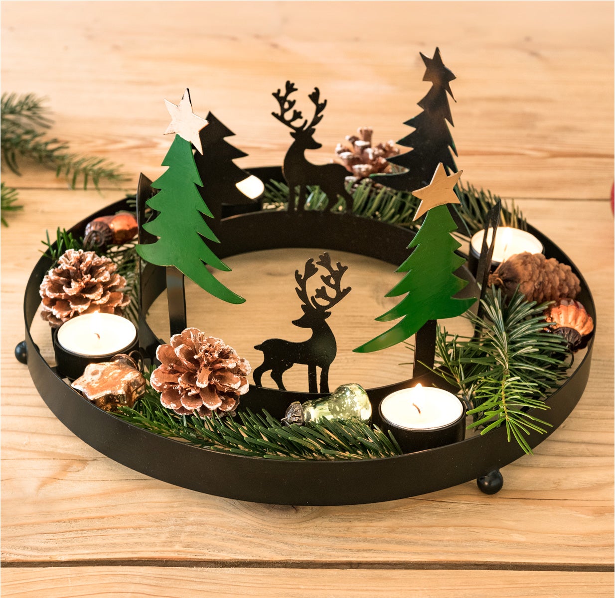 Adventskranz aus schwarzem Metall mit grünen Tannenbaumdekorationen, schwarzen Rentier-Silhouetten, Tannenzapfen, grünen Zweigen, kleinen gläsernen Christbaumkugeln und vier weißen Teelichtern auf Holzuntergrund