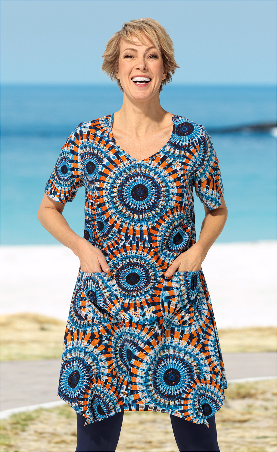 Frau trägt buntes Viskose-Kleid mit rundem Ausschnitt, kurzen Ärmeln und großen Taschen, Muster aus orange-blauen Kreisen, steht lächelnd am Strand mit blauem Himmel und Meer im Hintergrund