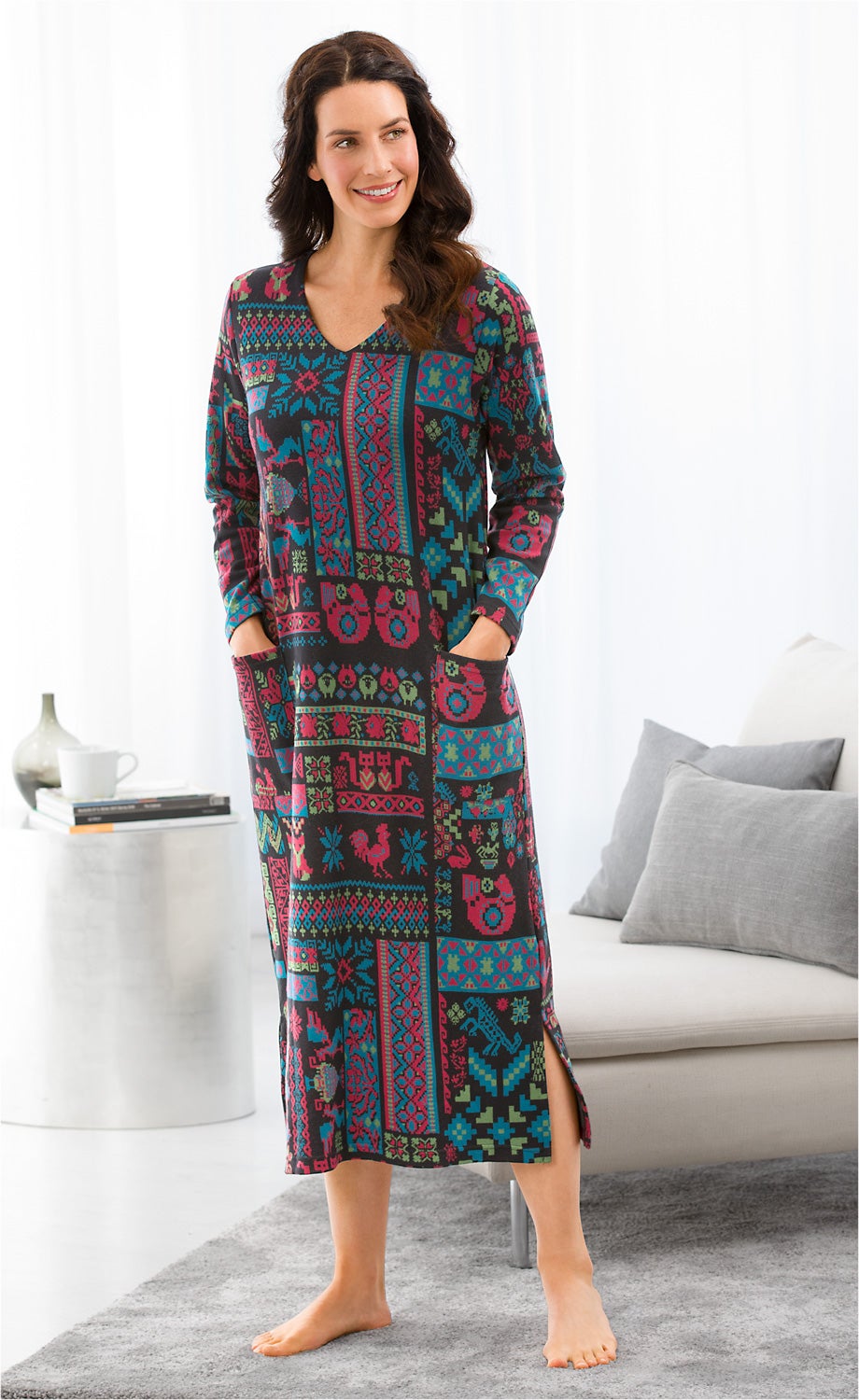 Frau trägt farbenfrohes, langärmeliges Baumwoll-Hauskleid mit geometrischem und floralen Muster, seitlicher Schlitz, steht barfuß im Wohnzimmer, Hände in den Taschen, lächelt, helle Gardinen und graues Sofa im Hintergrund.