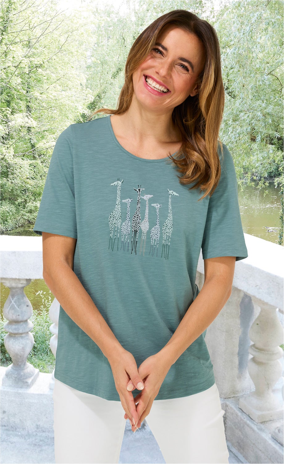 Frau trägt hellblaues Baumwoll-Shirt mit kurzärmeligem, rundem Ausschnitt und niedlichem Giraffen-Druck, steht lächelnd vor grünem Park mit Fluss und weißem Steinbalustraden-Geländer, trägt weiße Hose