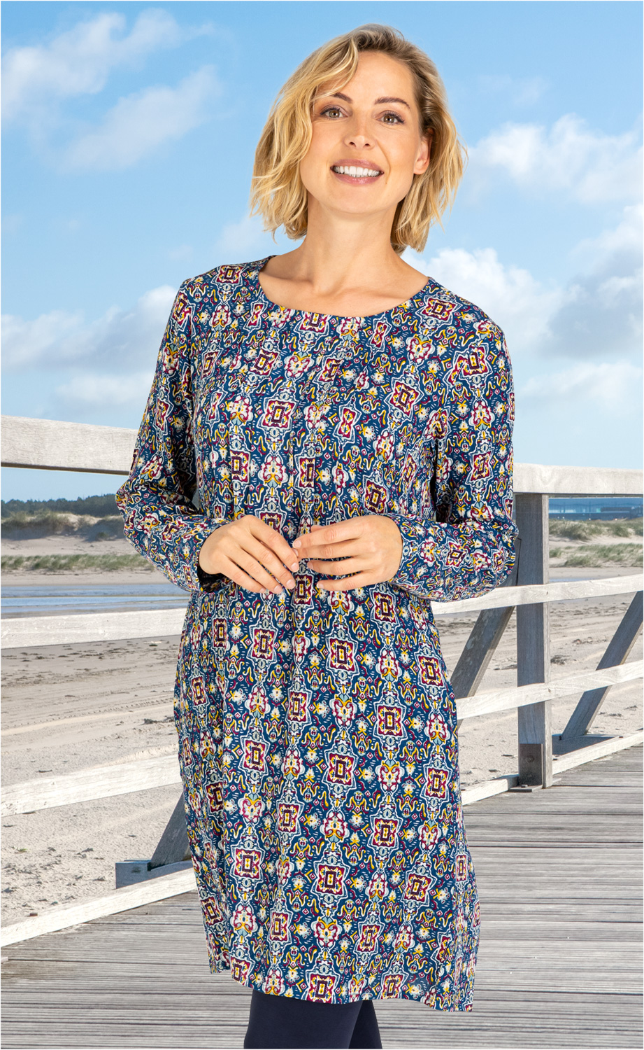 Frau trägt langärmeliges Viskose-Kleid mit buntem, geometrischem Muster in Blau, Gelb, Rot und Weiß, steht auf Holzsteg am Strand bei blauem Himmel, lächelt, blonde schulterlange Haare, legere Pose mit Händen vor dem Körper