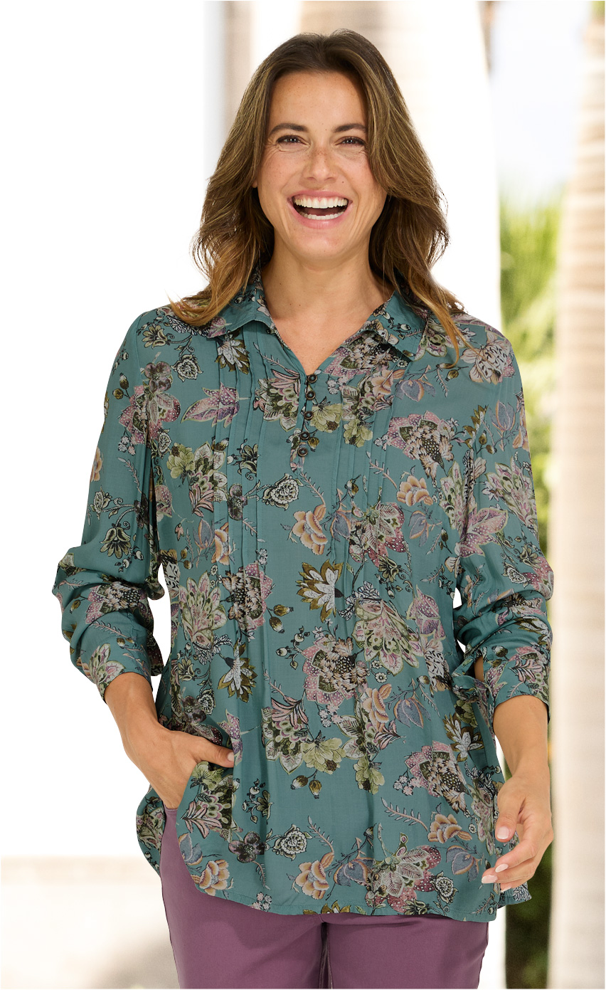 Frau trägt grüne Modal-Bluse mit floralem Muster, Langarm-Design, Knopfleiste und Hemdkragen, kombiniert mit lila Hose, lachend, Hände in Taschen, heller Hintergrund mit unscharfen Pflanzen und Säulen