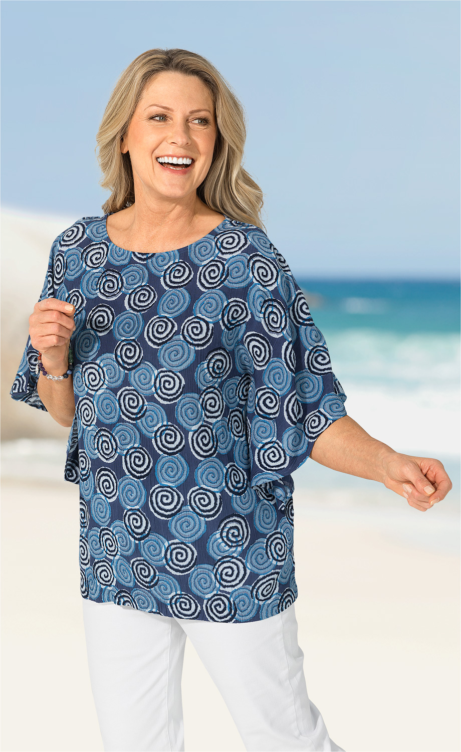 Frau in Viskose-Bluse mit blauem Spiralmuster, halblangen Ärmeln, steht lächelnd am Strand vor blauem Himmel und Meer, trägt weiße Hose und Armband, rechter Arm ausgestreckt