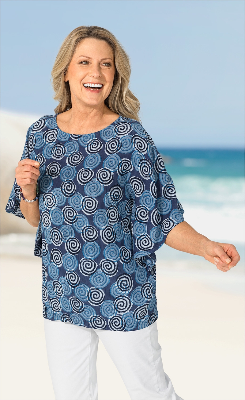 Frau in Viskose-Bluse mit blauem Spiralmuster, halblangen Ärmeln, steht lächelnd am Strand vor blauem Himmel und Meer, trägt weiße Hose und Armband, rechter Arm ausgestreckt