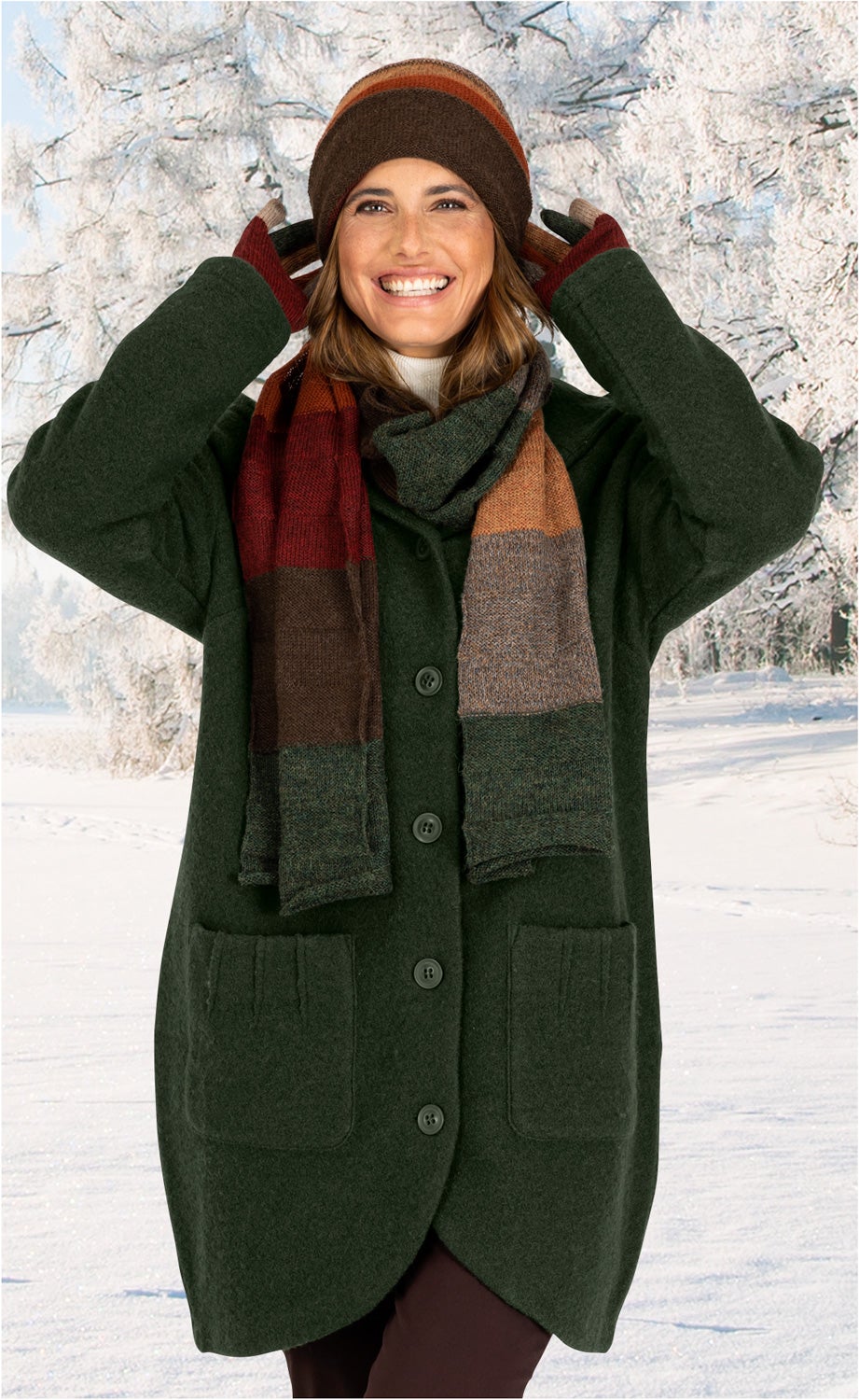 Frau trägt dunkelgrüne Woll-Jacke mit Knöpfen und aufgesetzten Taschen, bunte gestreifte Wollmütze, passende Wollhandschuhe und Schal, lächelt, steht vor verschneiter Winterlandschaft mit schneebedeckten Bäumen.