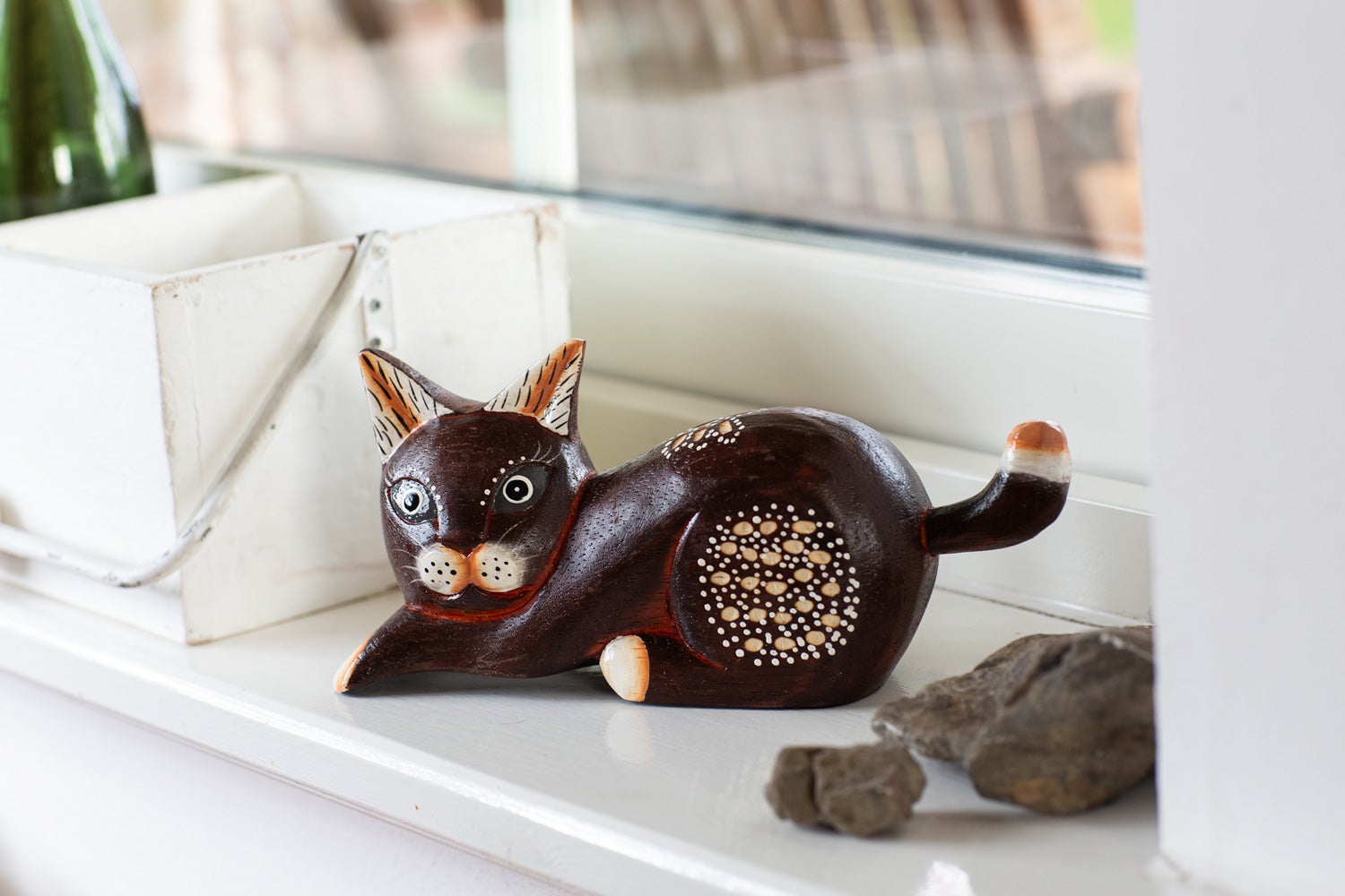 braune Deko-Katzenfigur aus Holz mit weißen Punkten und schwarzen Details, liegend auf weißem Fensterbrett neben weißem Holzkasten und Steinen, im Hintergrund unscharfes Fenster und Gartenbereich