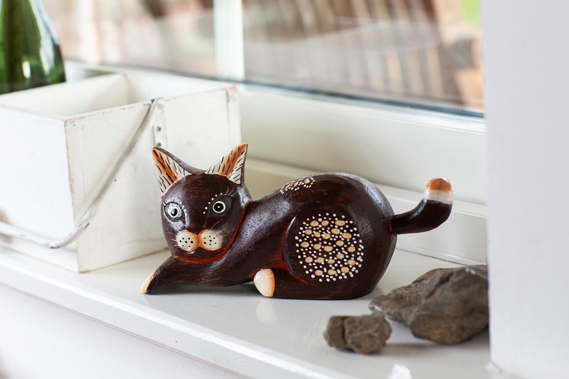 braune Deko-Katzenfigur aus Holz mit weißen Punkten und schwarzen Details, liegend auf weißem Fensterbrett neben weißem Holzkasten und Steinen, im Hintergrund unscharfes Fenster und Gartenbereich