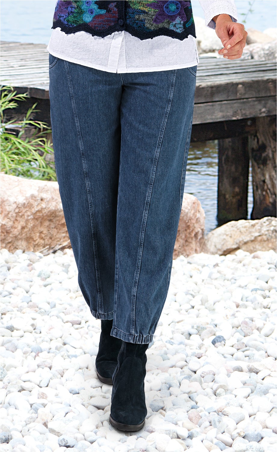 Dunkelblaue Baumwoll-Hose mit elastischen Bündchen, getragen von Person mit weißer Bluse und bunter Strickweste, dazu schwarze Stiefeletten, stehend auf hellem Kieselsteinboden vor einem hölzernen Steg und Wasser im Hintergrund