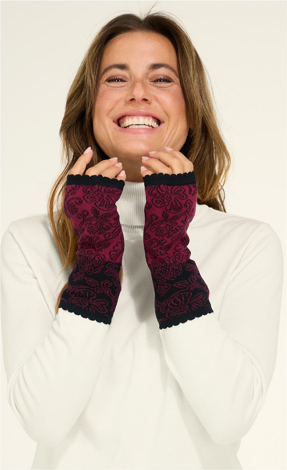 lächelnde Frau trägt weiße Rollkragenpullover und bordeauxrote Baumwoll-Pulswärmer mit schwarzem Blumenmuster, Finger frei und mit gewelltem Saum