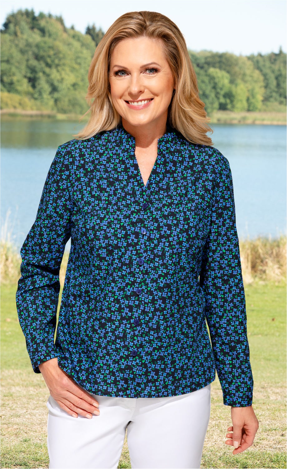 Frau mit blonder mittellanger Frisur trägt dunkelblaue Baumwoll-Bluse mit kleinem blauen und grünen Blumenmuster, steht vor See und Wald, lächelt, kombiniert mit weißen Hosen, rechte Hand in Hosentasche, linke Hand locker seitlich