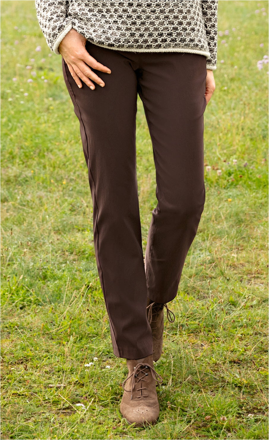 Beine in brauner Viskose-Mix-Hose und beigen Schnürschuhen auf Grasfläche, Oberteil mit gemustertem Strickpullover, eine Hand auf Oberschenkel, natürliche Outdoor-Umgebung, modische legere Herbstbekleidung
