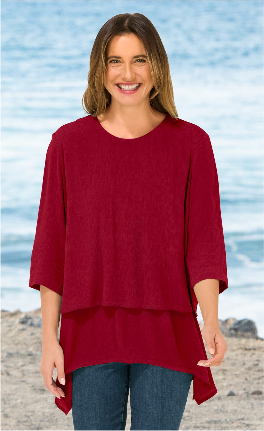 Frau trägt rote Viskose-Tunika mit 3/4-Ärmeln und asymmetrischem Saum, lächelt, steht am Strand mit Meer im Hintergrund, darunter blaue Jeans