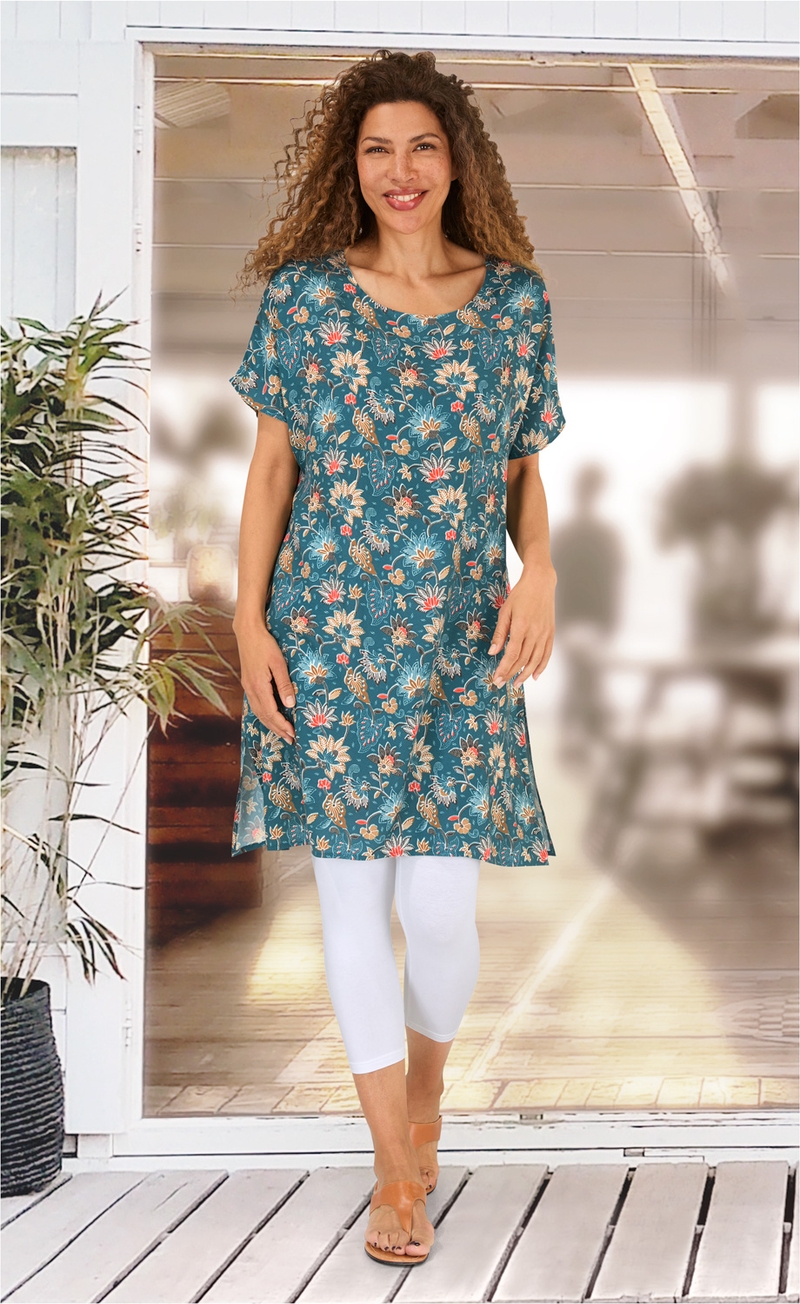 Frau trägt blauen Viskose-Kaftan mit floralem Muster, weiße Leggings und braune Sandalen, steht vor einer Glas-Schiebetür mit Pflanzen im Hintergrund, lächelt, lange lockige Haare, helle Wohnraumbeleuchtung