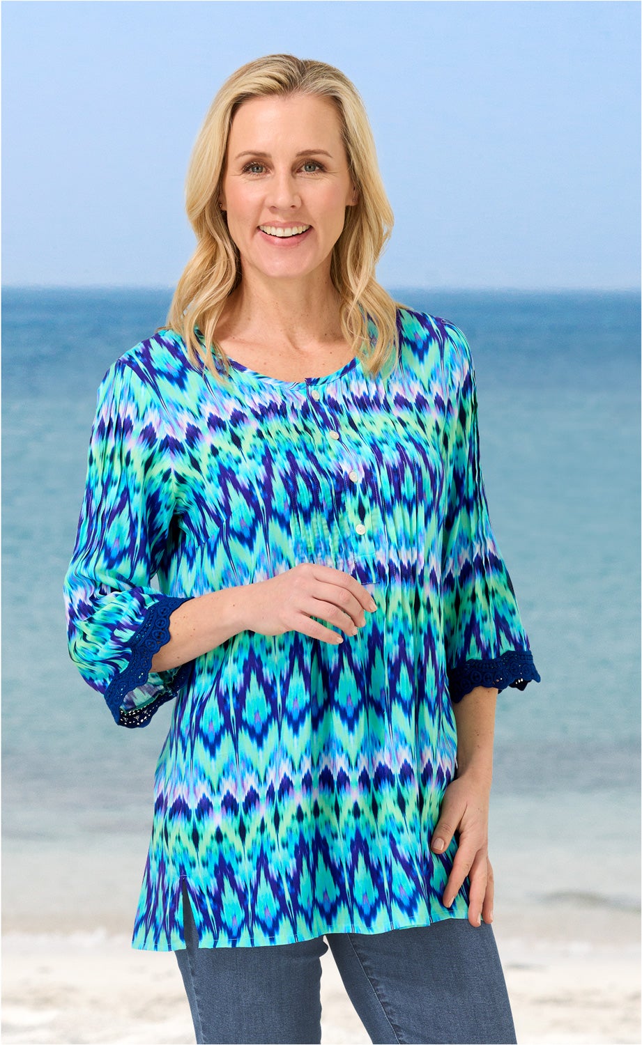 Frau am Strand trägt bunte Modal-Bluse in Blau- und Grüntönen mit 3/4-Ärmeln und Knopfleiste, blaue Jeans, lächelt, hellblonde lange Haare, Meereskulisse im Hintergrund