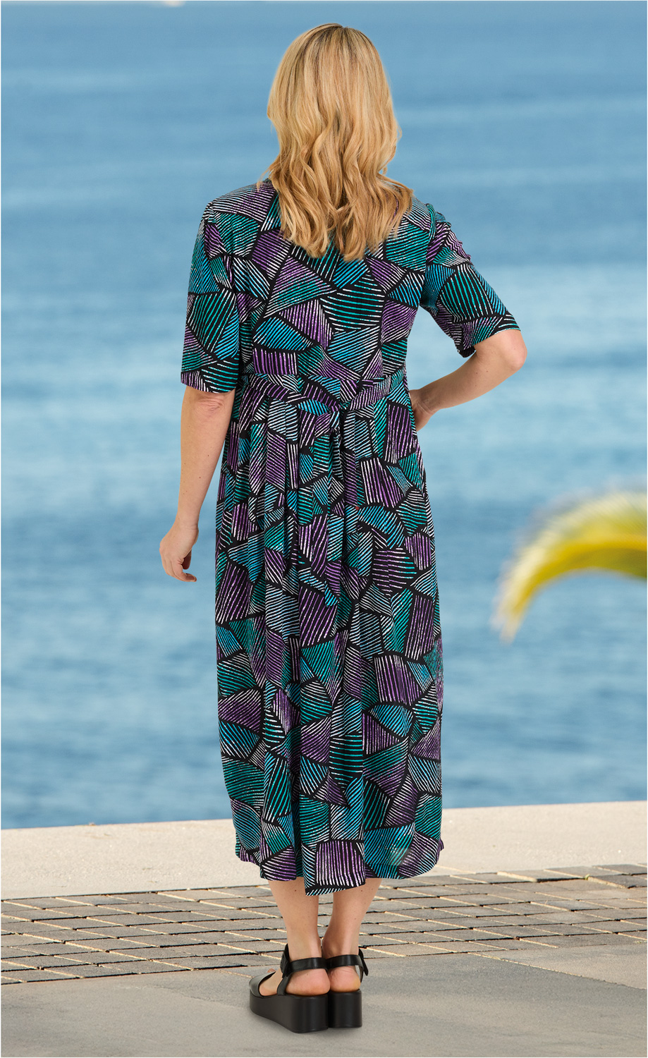 Frau von hinten in einem bunten Viskose-Kleid mit abstraktem Muster in Türkis, Lila und Schwarz, halblange Ärmel, schwarze Sandalen, stehend an einem Wasserufer mit blauem Meer im Hintergrund
