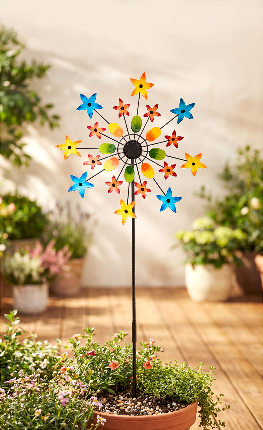 Bunter Deko-Gartenstecker mit rot-gelben, blauen und grünen Blumenmotiven auf schwarzem Stab, steckend in einem Blumentopf mit kleinen blühenden Pflanzen auf Holzterrasse, unscharfer Gartenhintergrund