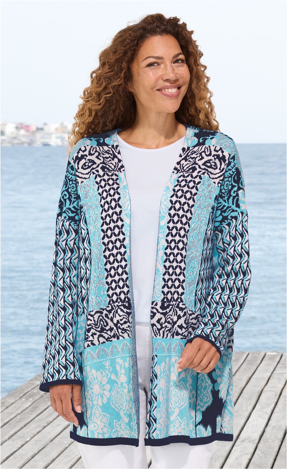 Frau trägt offene Baumwoll-Strickjacke in Blau- und Weißtönen mit floralem und geometrischem Muster, weißes Shirt, weiße Hose, stehend auf Holzsteg am Meer, lockige Haare, lächelnd