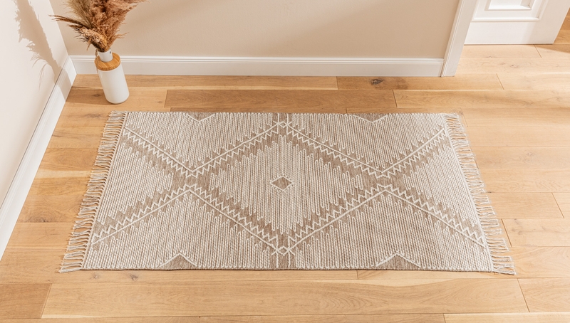 Teppich Mu.natur 70x140 auf hellem Holzboden, beige mit weißem, geometrischem Muster und Zickzacklinien, Fransen an den kurzen Seiten, im Hintergrund helle Wand mit einer weißen Vase und getrockneten Gräsern