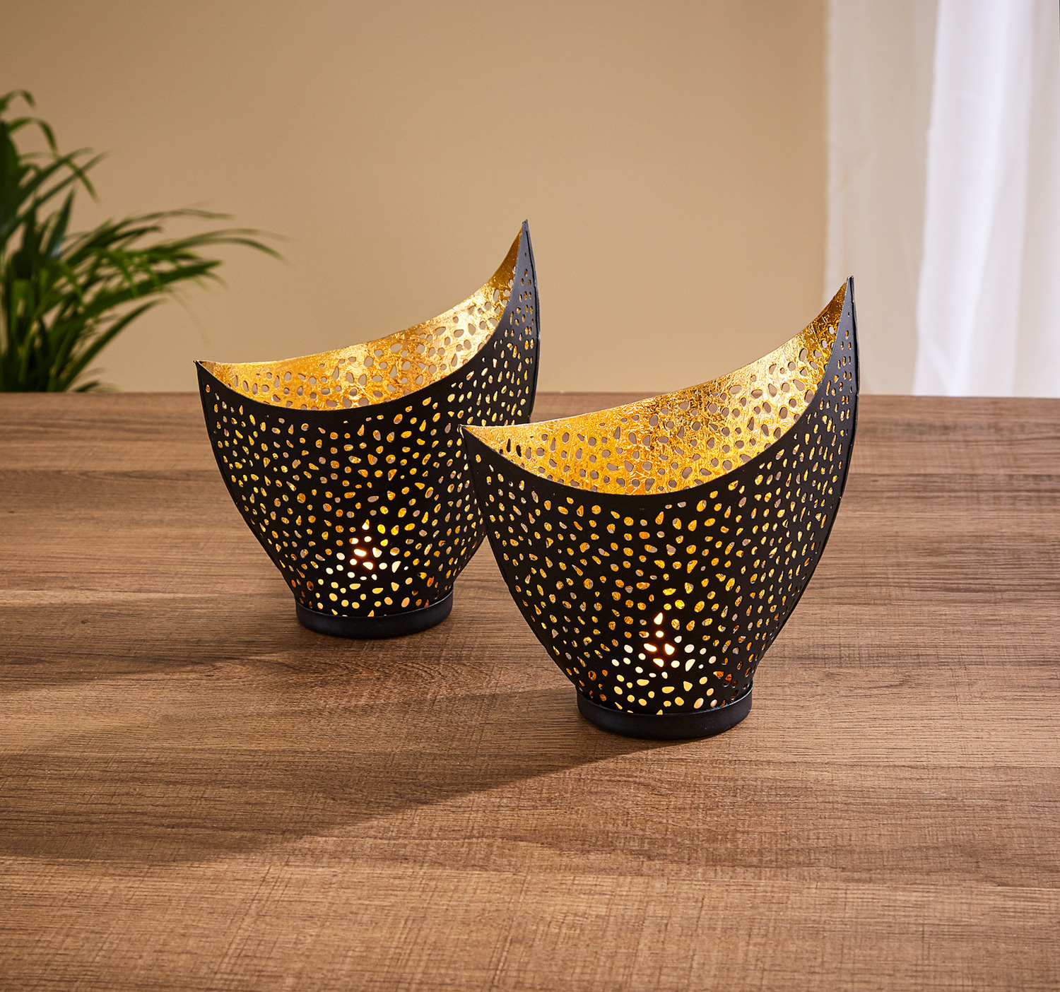 Zweier-Set schwarzer Windlichter mit goldener Innenseite, perforiertem Muster und asymmetrischer Form, beleuchtet, auf wooden Tisch, Hintergrund mit grüner Pflanze und hellem Vorhang