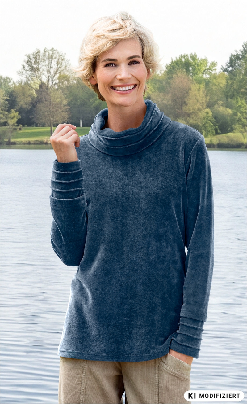 lächelnde Frau mit kurzem blondem Haar, trägt blaues Langarmshirt mit weitem Kragen, steht am See mit Bäumen im Hintergrund, linke Hand hebt sie leicht, rechte Hand in Tasche beige Hose